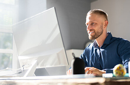 Ein Mann im blauen Hemd vor einem Desktopcomputer, Hintergrund scheint die Sonne durch das Fenster